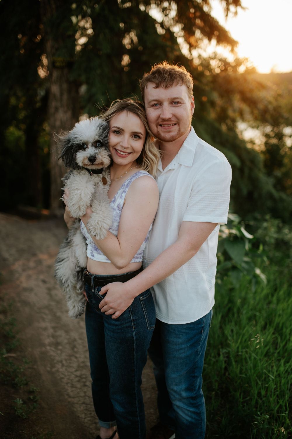 A couple poses with their small fluffy white dog during golden hour on a forest trail at sunset.