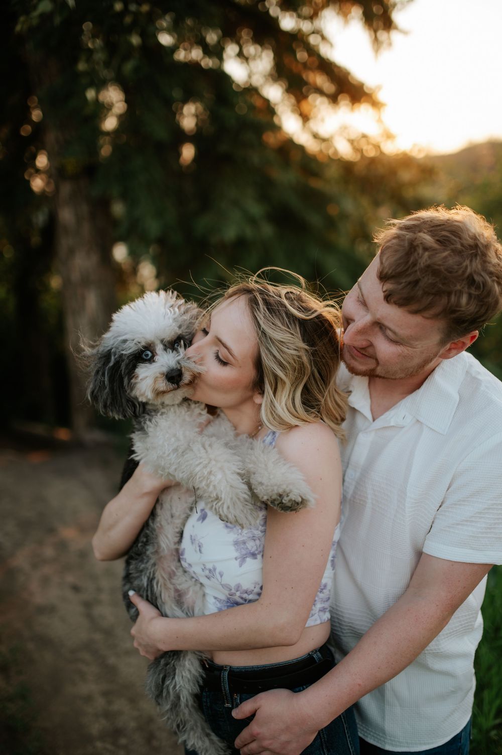 Loving couple embraces their small fluffy white and gray dog outdoors during golden sunset.