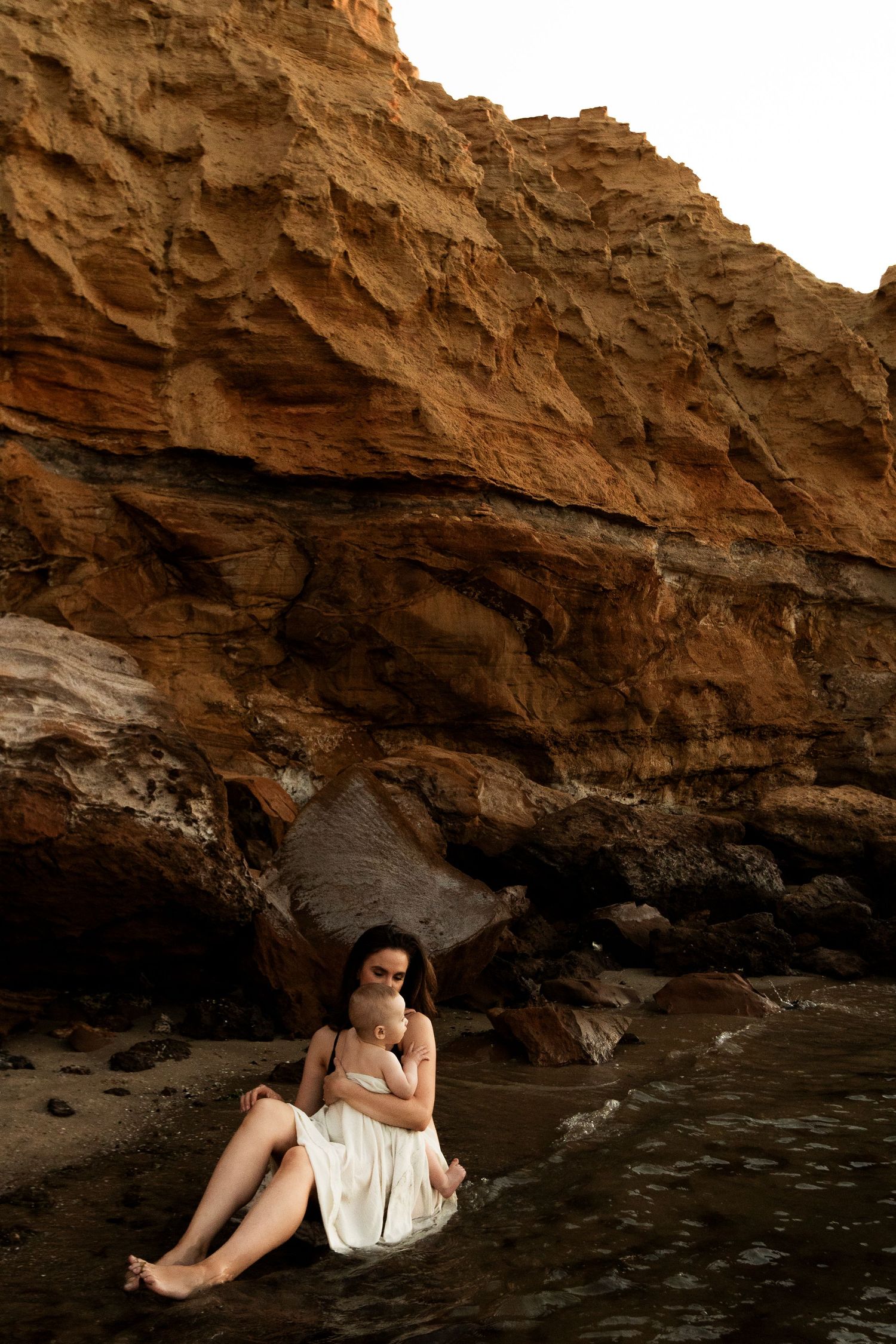 A woman in white dress sits contemplatively under dramatic rocky cliffs by the ocean.