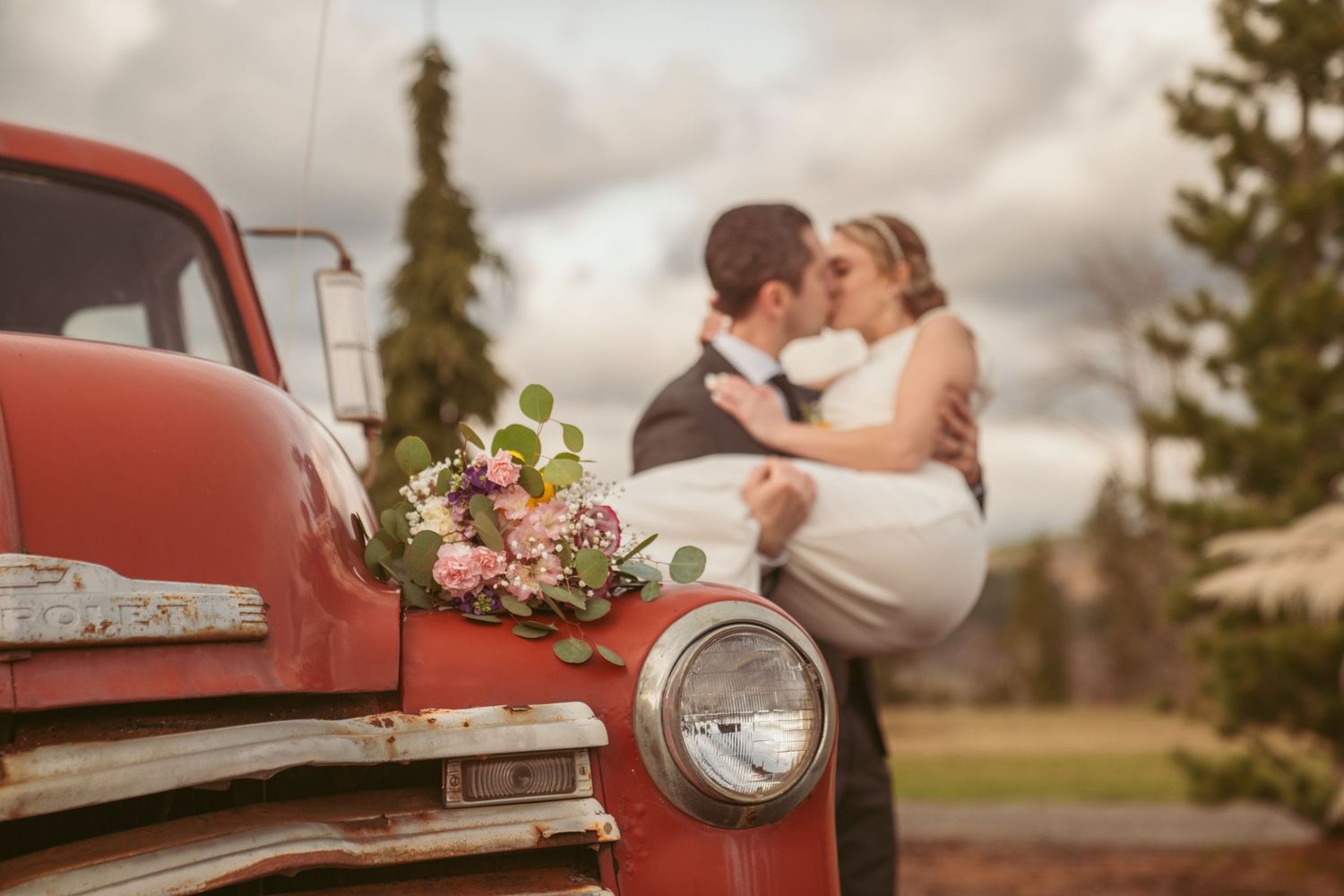 A couple stands in a sunlit meadow wearing wedding attire surrounded by evergreen trees during golden hour.