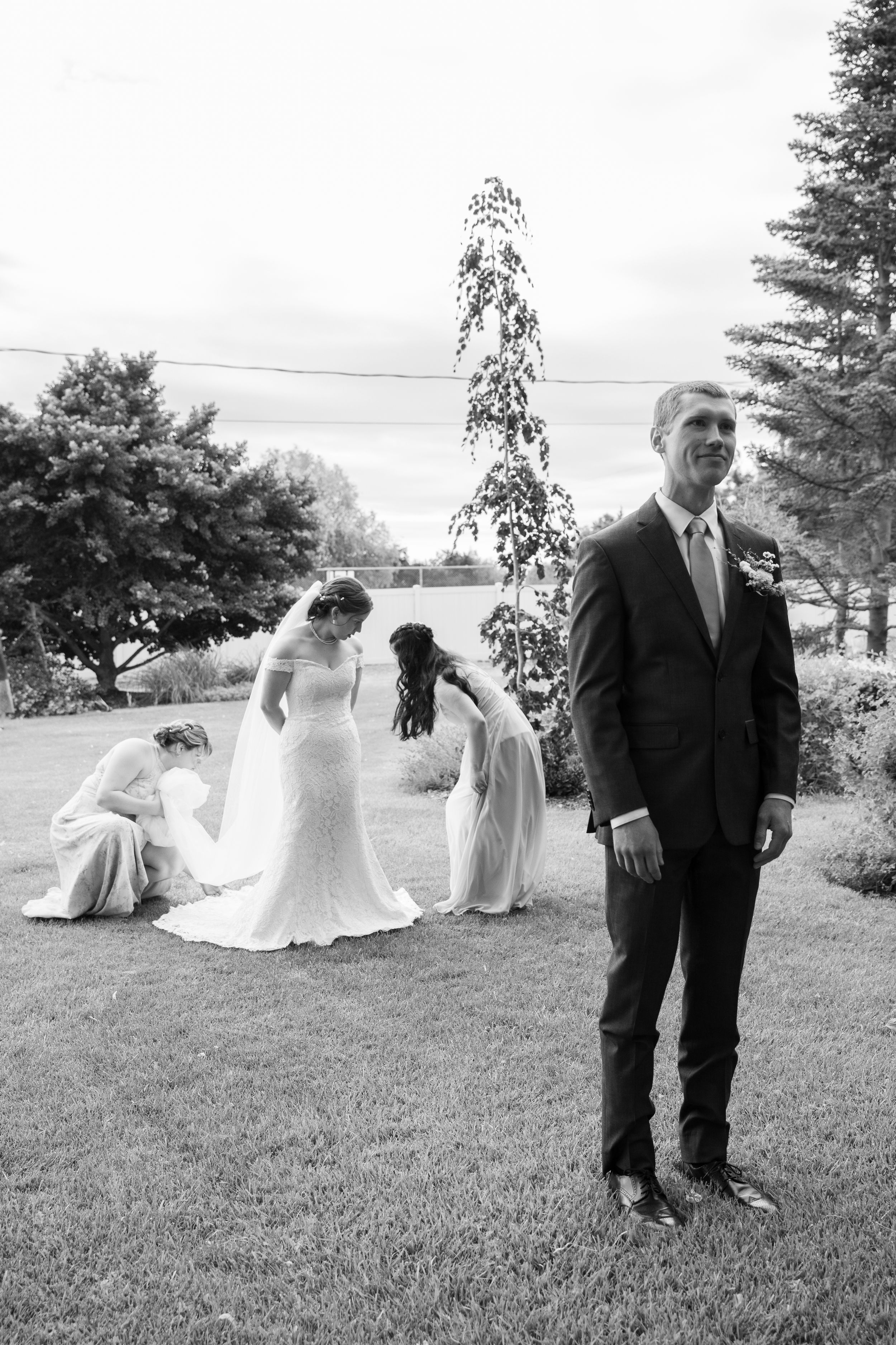 Black and white wedding photo showing a suited groom standing while the bridal party gathers in the garden background.