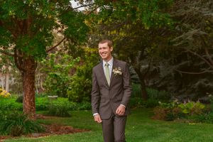 A person in a brown suit stands in a lush garden setting with trees and greenery in the background.