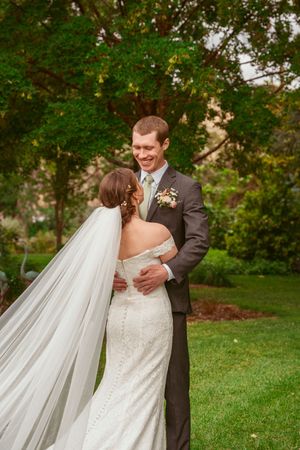 A flowing white wedding veil catches the breeze as a couple embraces in a lush garden setting during their special day.