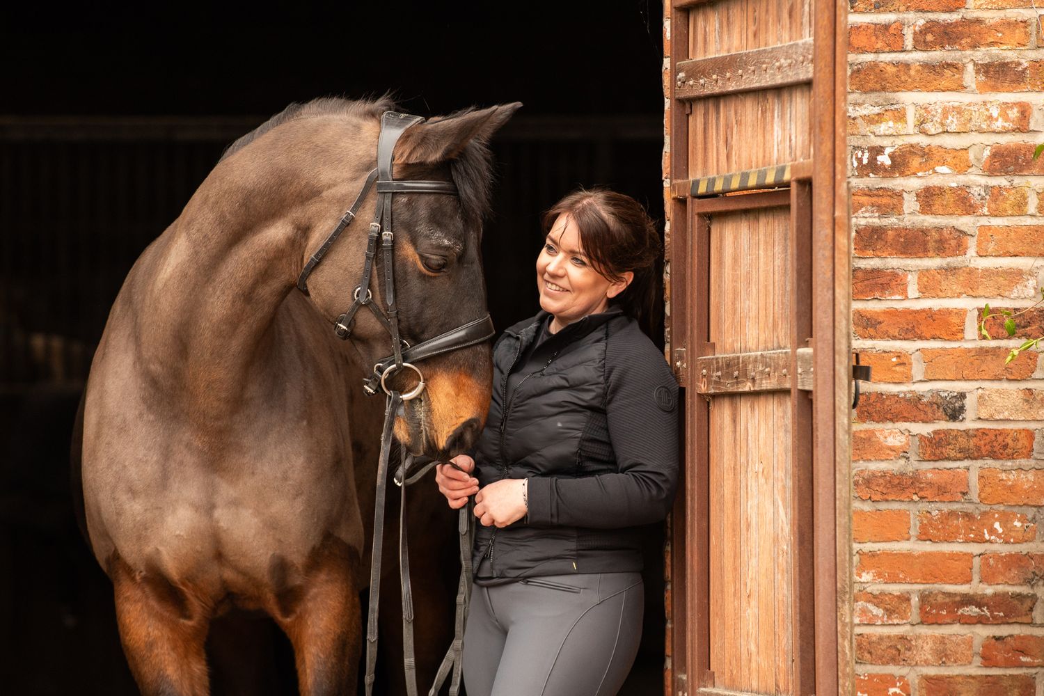 Person in black jacket grooms a tall brown horse in a brick stable with wooden doors.