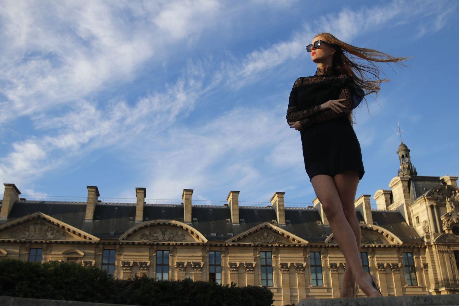 A model stands on the right side of the image, with the iconic buildings of the Louvre Museum elegantly displayed in the background.