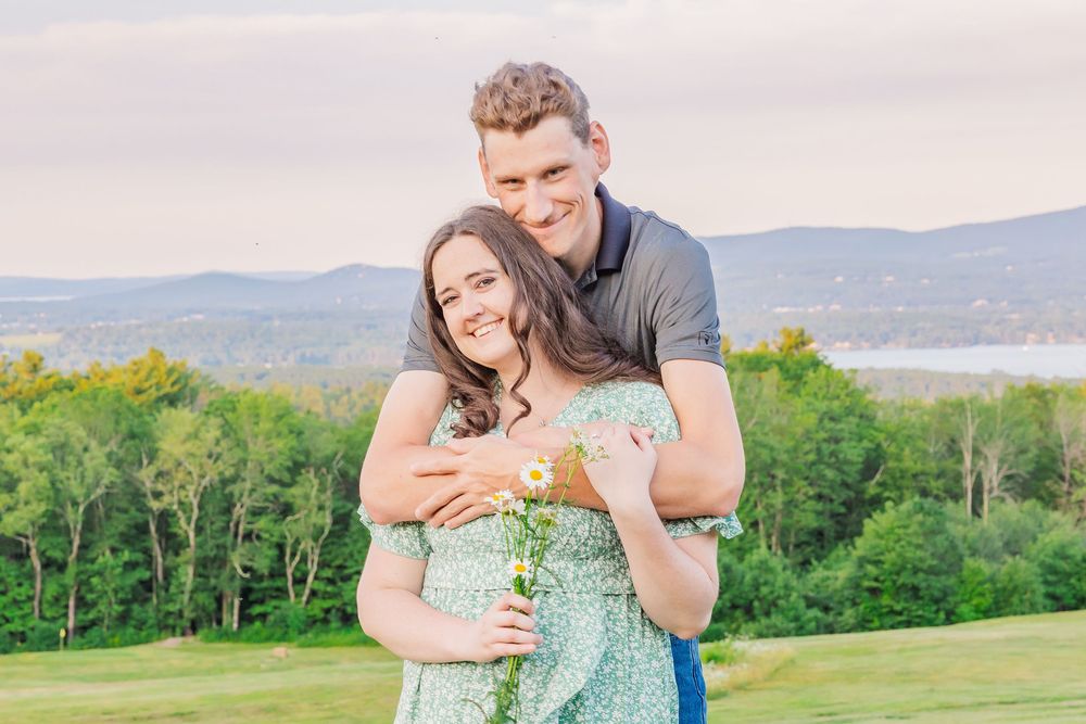A couple embraces on a hilltop overlooking mountains and a lake on a sunny summer day.