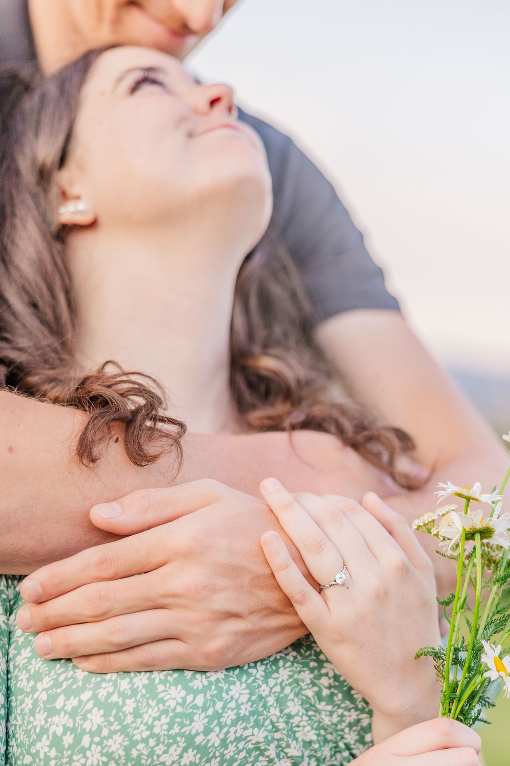 A woman wearing a green floral dress lying down showing off her engagement ring with white flowers visible.