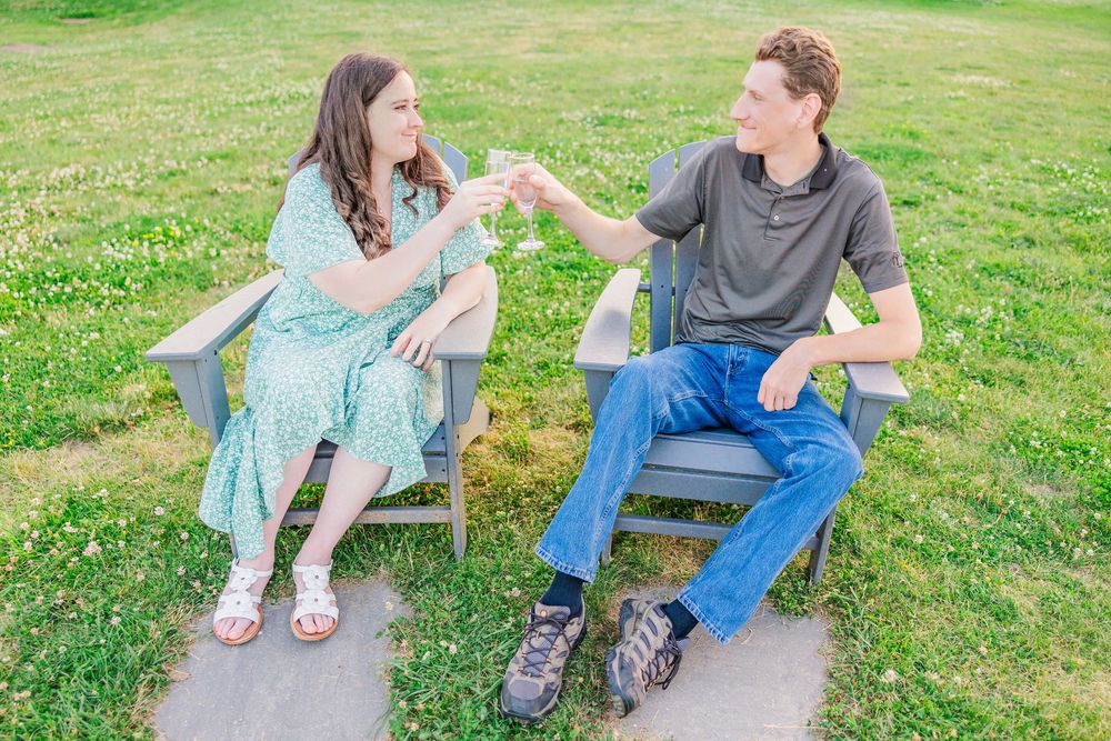 A couple sits on a wooden bench in a grassy field sharing casual conversation on a sunny day.