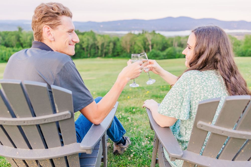 Two people sit in Adirondack chairs clinking wine glasses while enjoying a scenic mountain view on a summer evening.