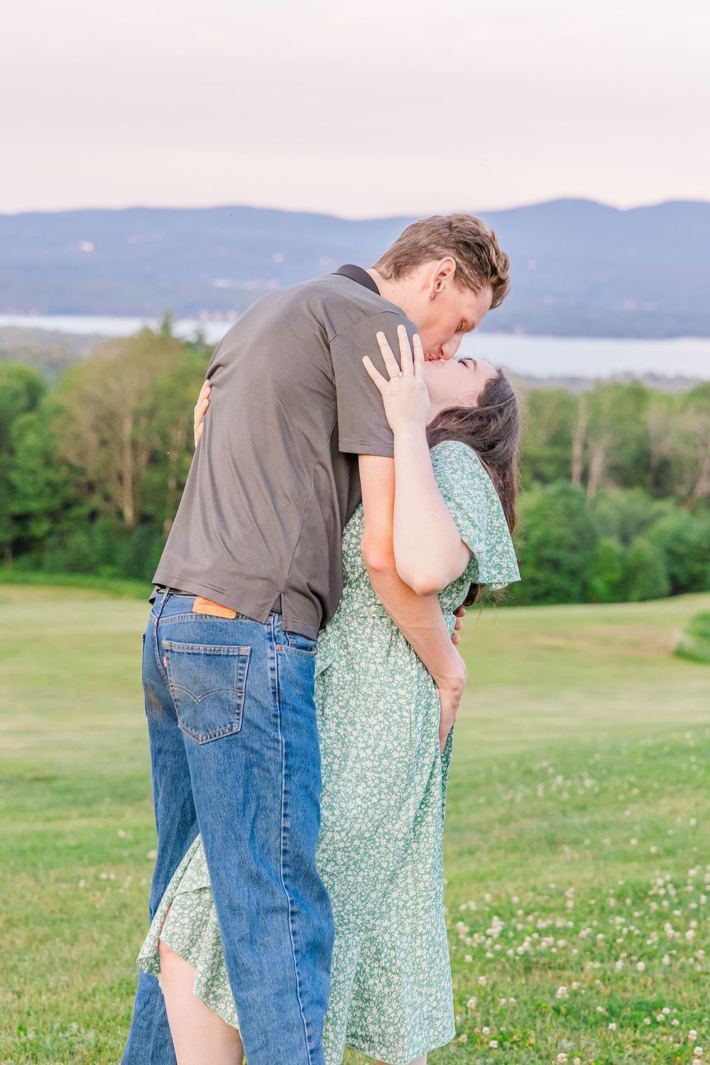 A romantic moment shared between a couple in a green field with mountains and lake in the background.