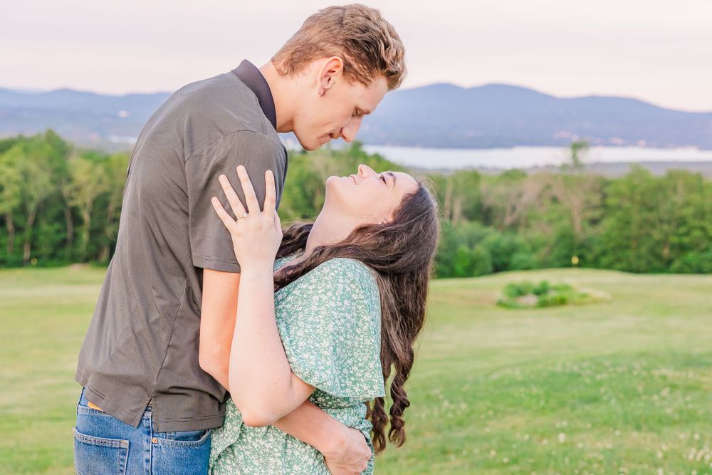 A couple shares a romantic embrace against a scenic mountain backdrop on a grassy hillside.