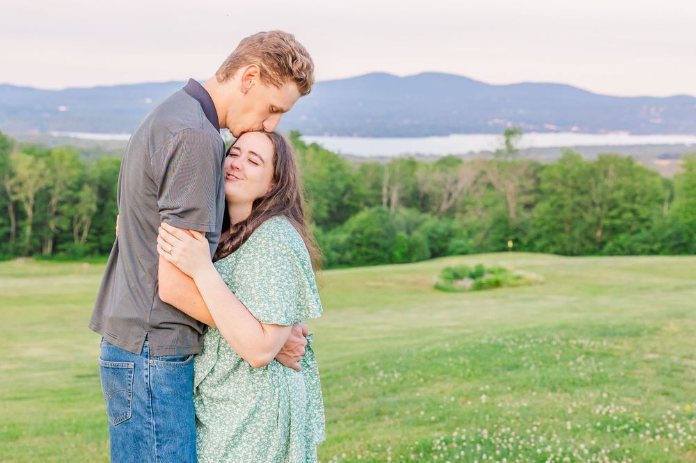 A romantic couple stands in a scenic meadow surrounded by mountains and greenery during golden hour.