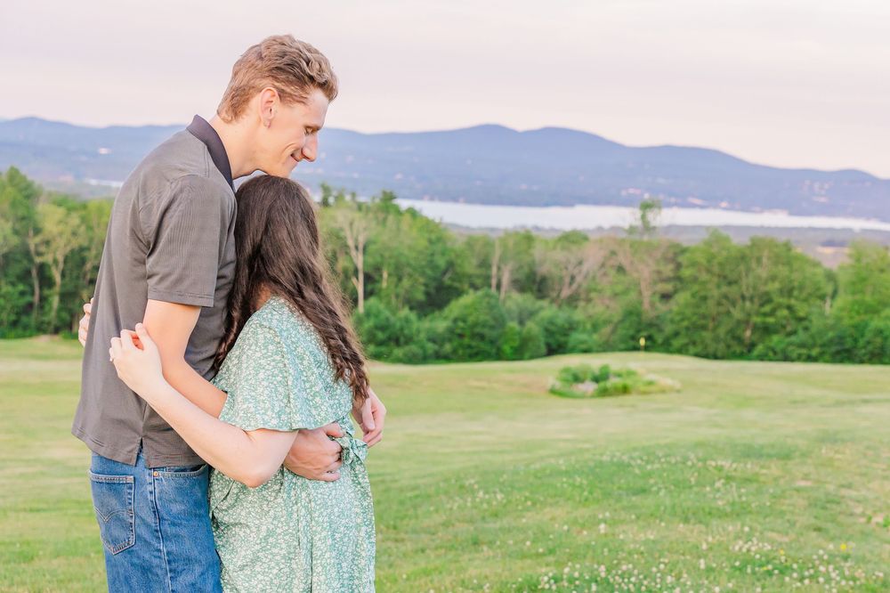 A couple embraces while standing on a grassy hillside overlooking a lake and mountains in the distance.