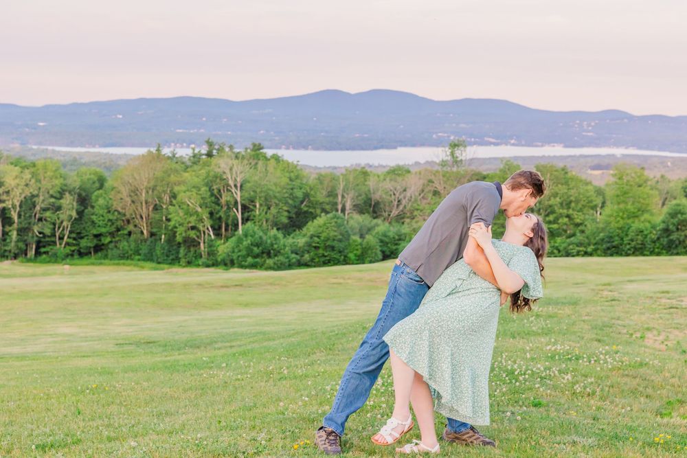 A couple embraces in a flowing dress against rolling hills and mountain landscape during sunset.