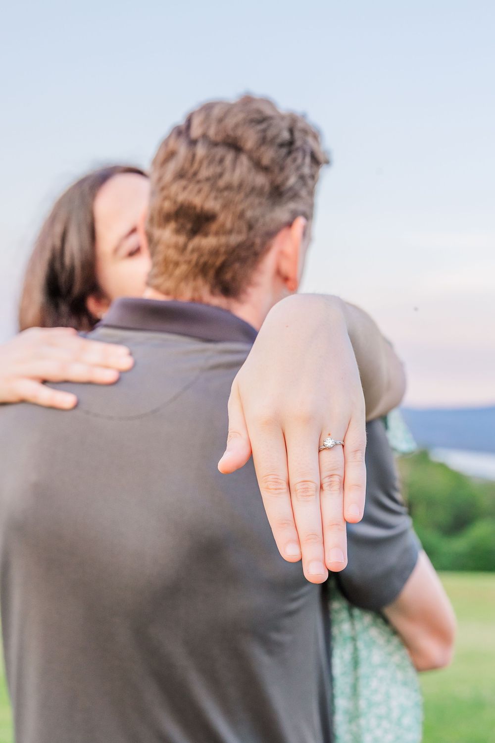 A newly engaged couple embraces outdoors while showing off the diamond engagement ring on her extended hand.