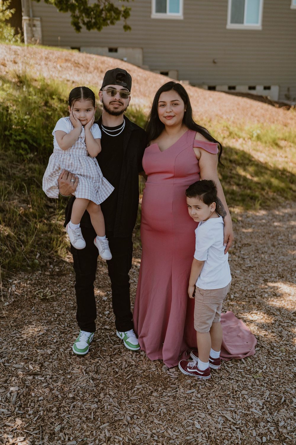Family of four posing outdoors with pregnant mother in a pink maxi dress.