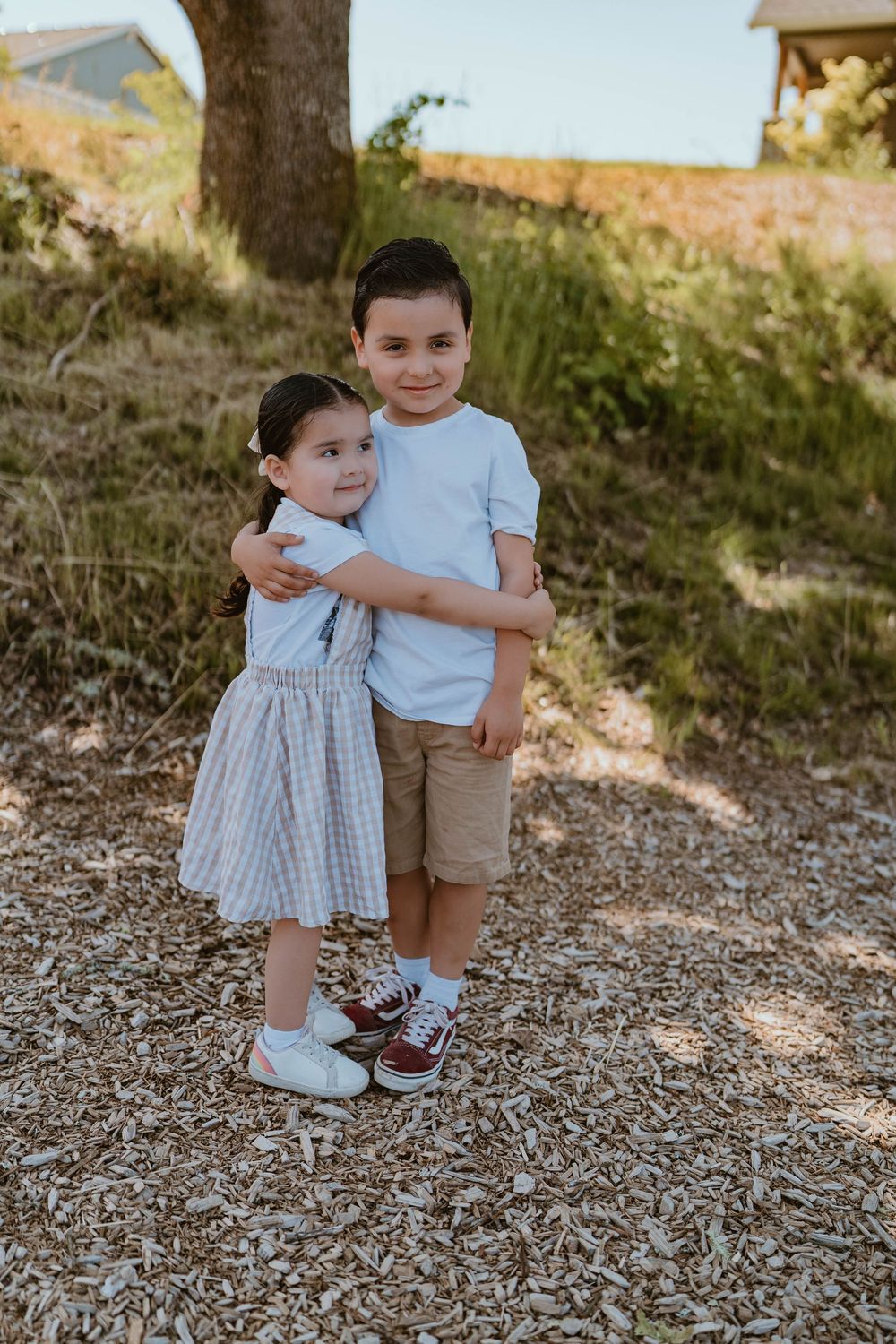 Two young siblings hugging outdoors, boy in white tee and girl in plaid dress.