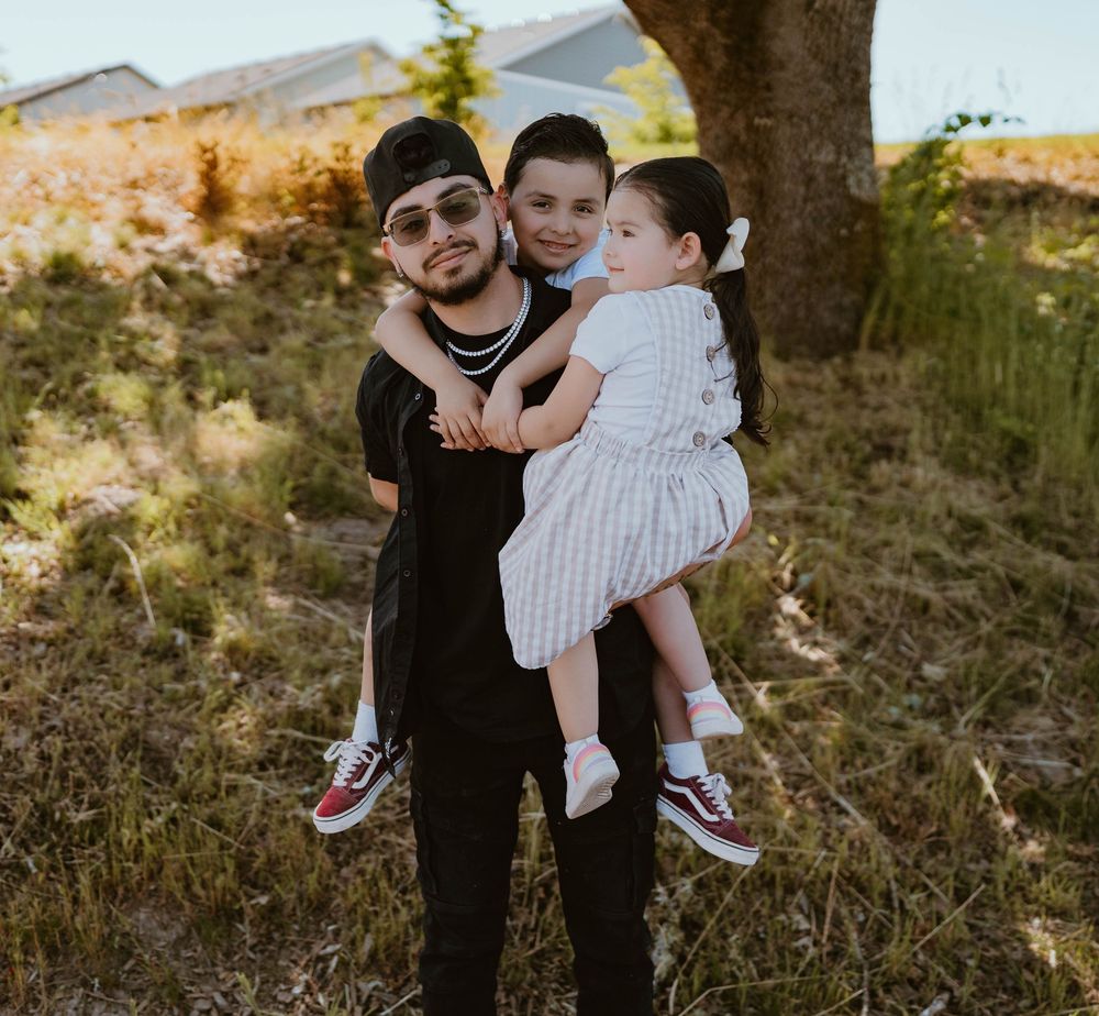 Father holding two young children in his arms, smiling outdoors near a tree.