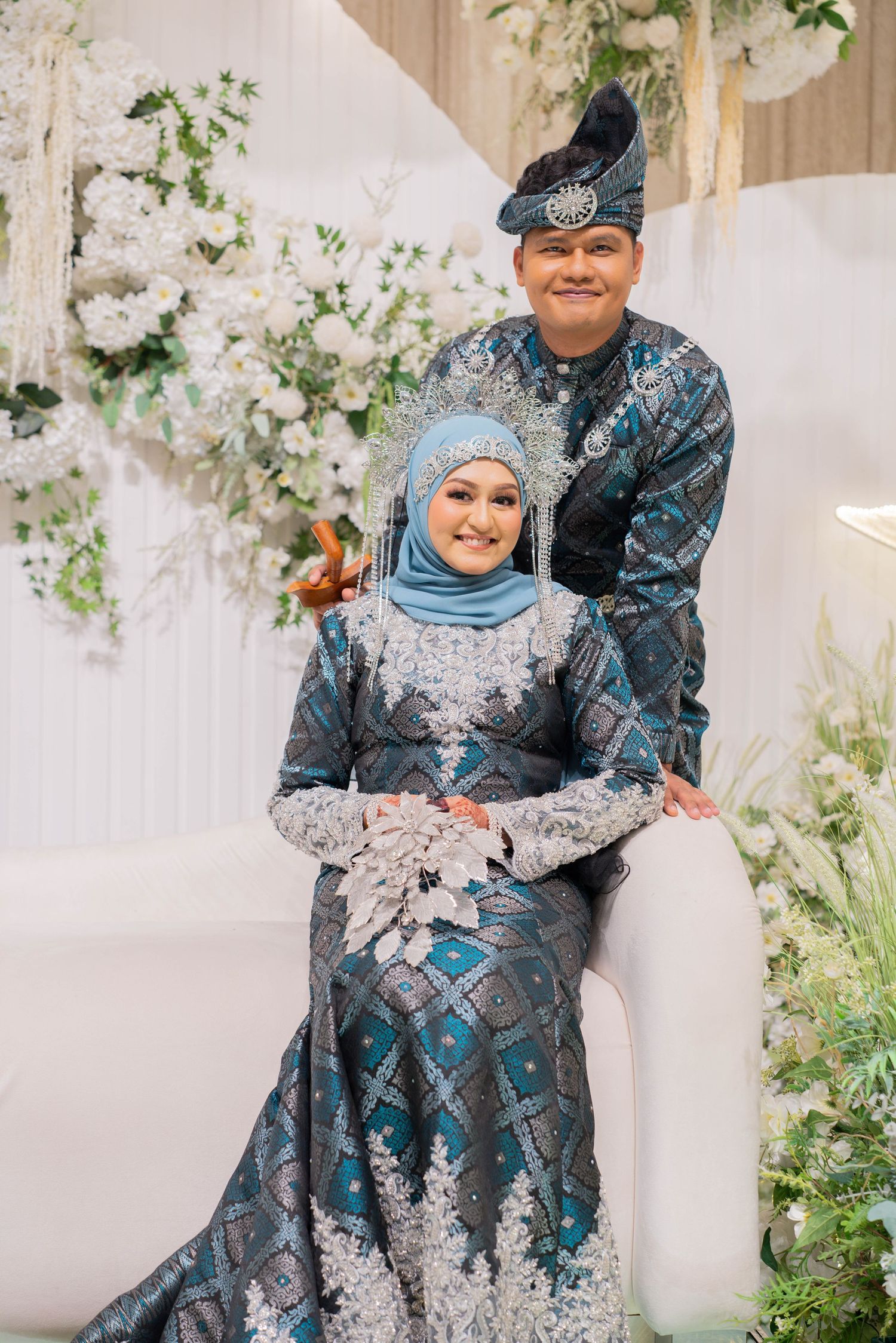 A couple wearing traditional blue and silver formal attire poses together among white floral decorations at a special event.