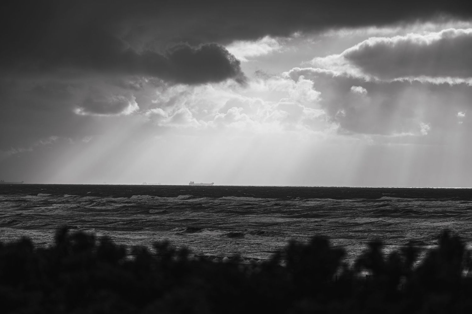 Dramatic sunbeams pierce through dark clouds over a turbulent ocean in black and white.