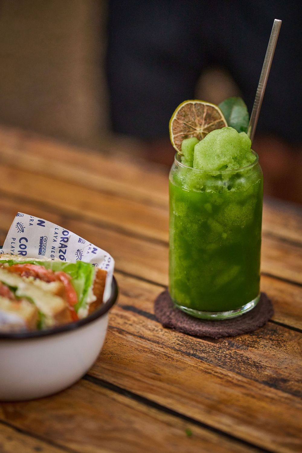 A vibrant green frozen cocktail garnished with lime sits on a rustic wooden table next to a small appetizer bowl.