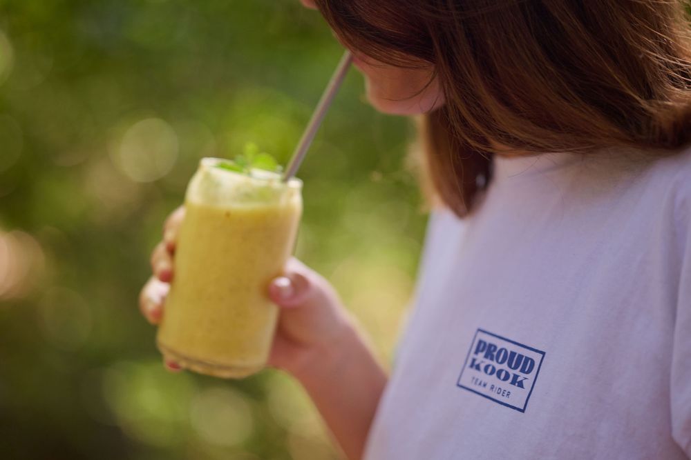 A yellow smoothie in a clear glass is held up in front of blurred green outdoor scenery.