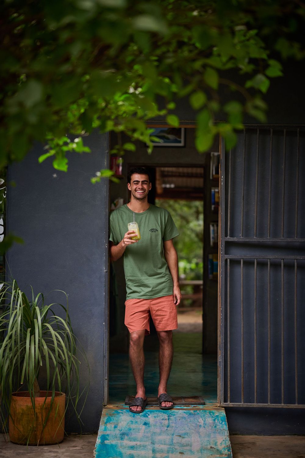 Someone stands in a doorway wearing a green t-shirt and coral shorts while holding a drink on a summer day.