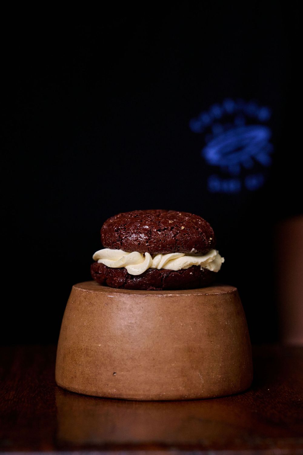 Chocolate whoopie pie dessert with cream filling sits atop a wooden pedestal against a dark background with blue lighting.