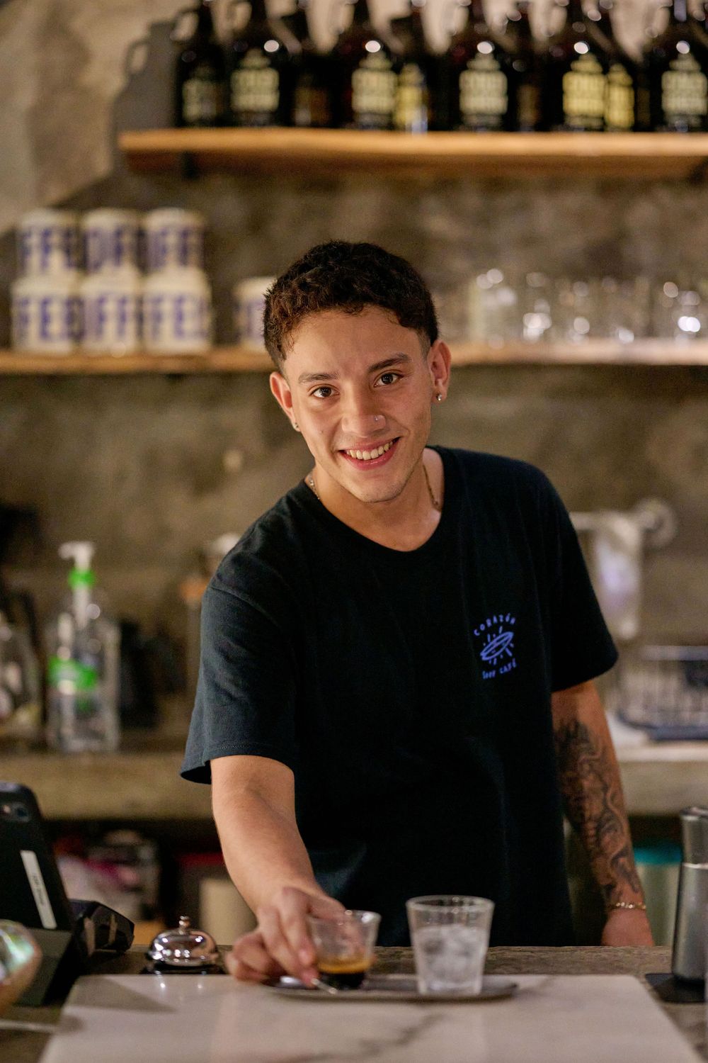 Bartender in black t-shirt smiles while working behind a bar counter with glasses and bottles visible in background.