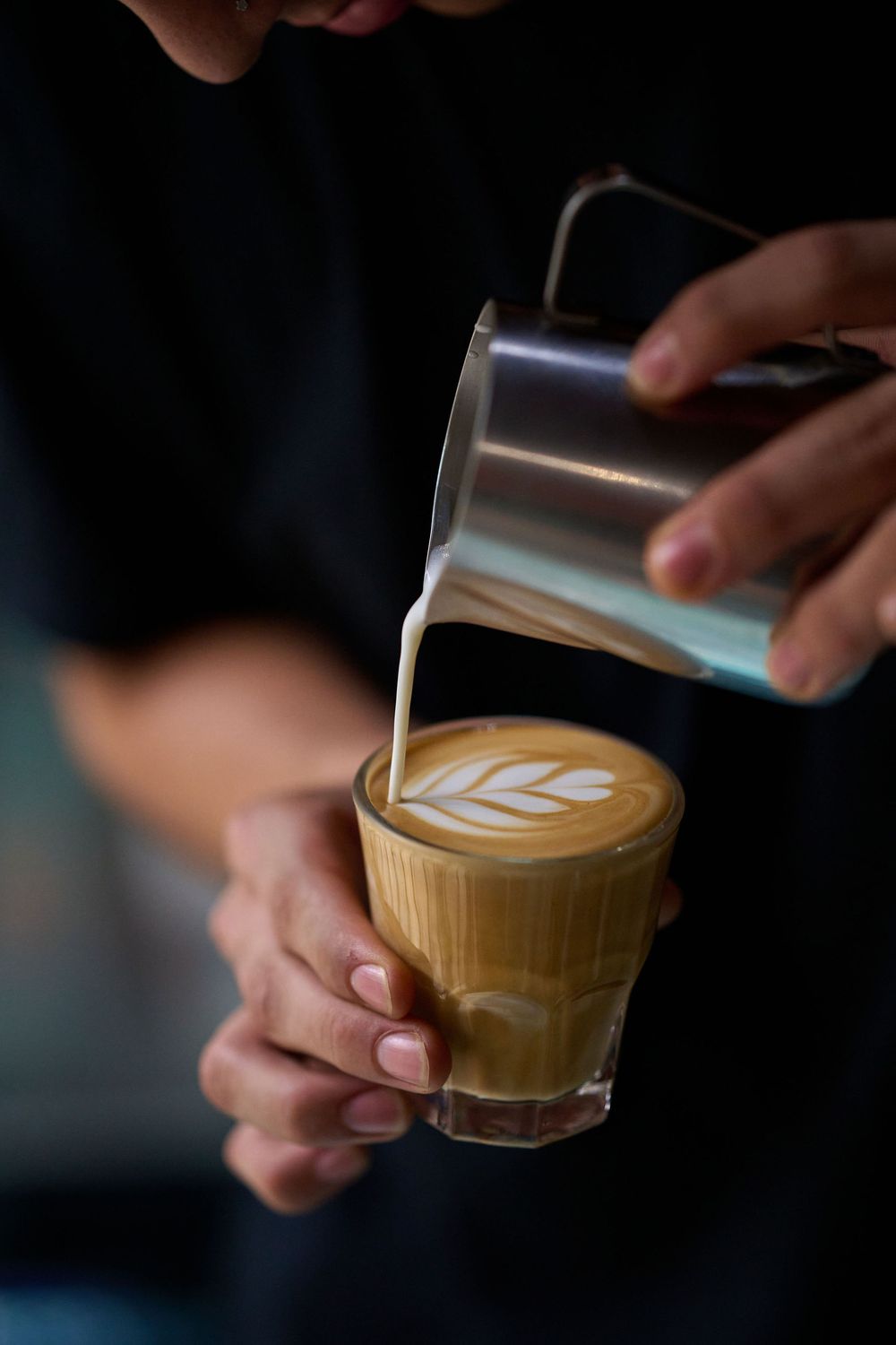 A barista pours steamed milk into a glass of coffee creating a delicate latte art leaf pattern.