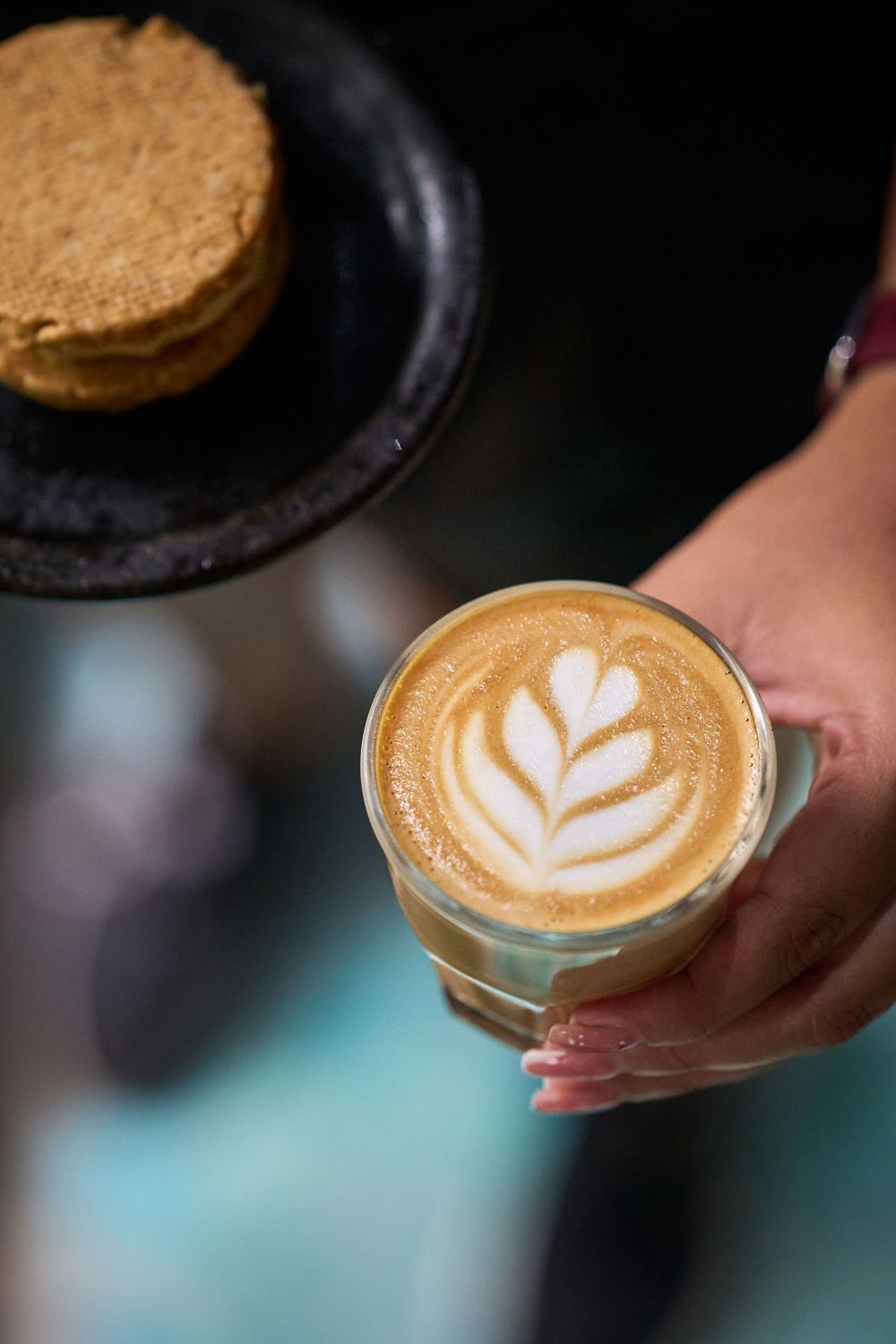 A cappuccino with latte art showing a delicate leaf pattern sits alongside a cookie on a dark plate.