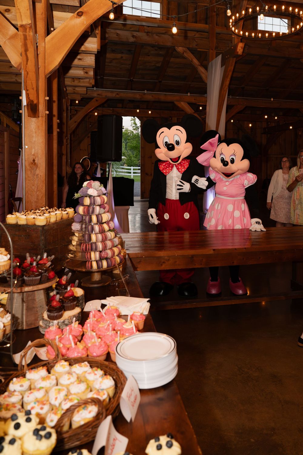 Mickey and Minnie Mouse characters stand behind a dessert display at a rustic barn venue with string lights and wooden beams.