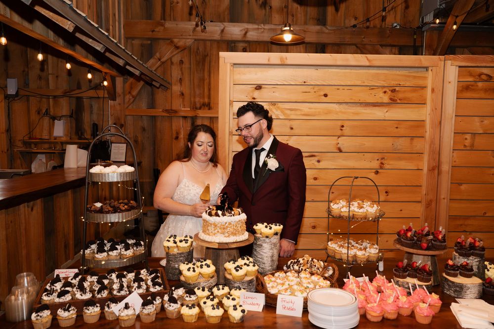 Wedding couple stands beside rustic dessert table with tiered cake and cupcakes in wooden barn setting.