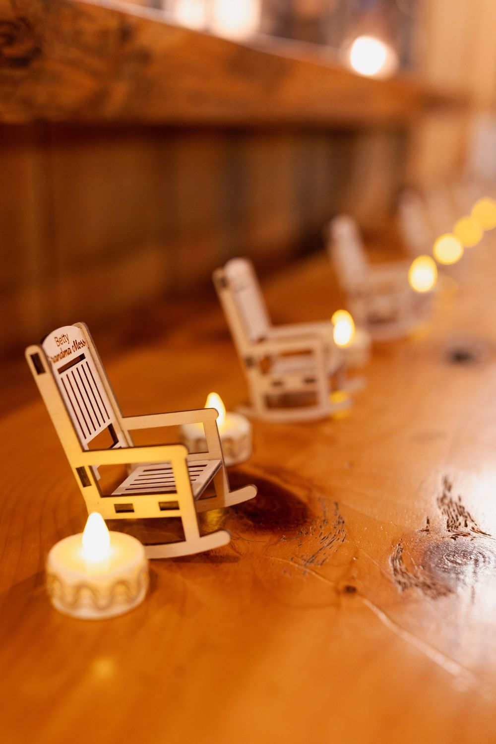 Miniature white rocking chairs and tea light candles arranged on wooden surface with soft bokeh lighting.