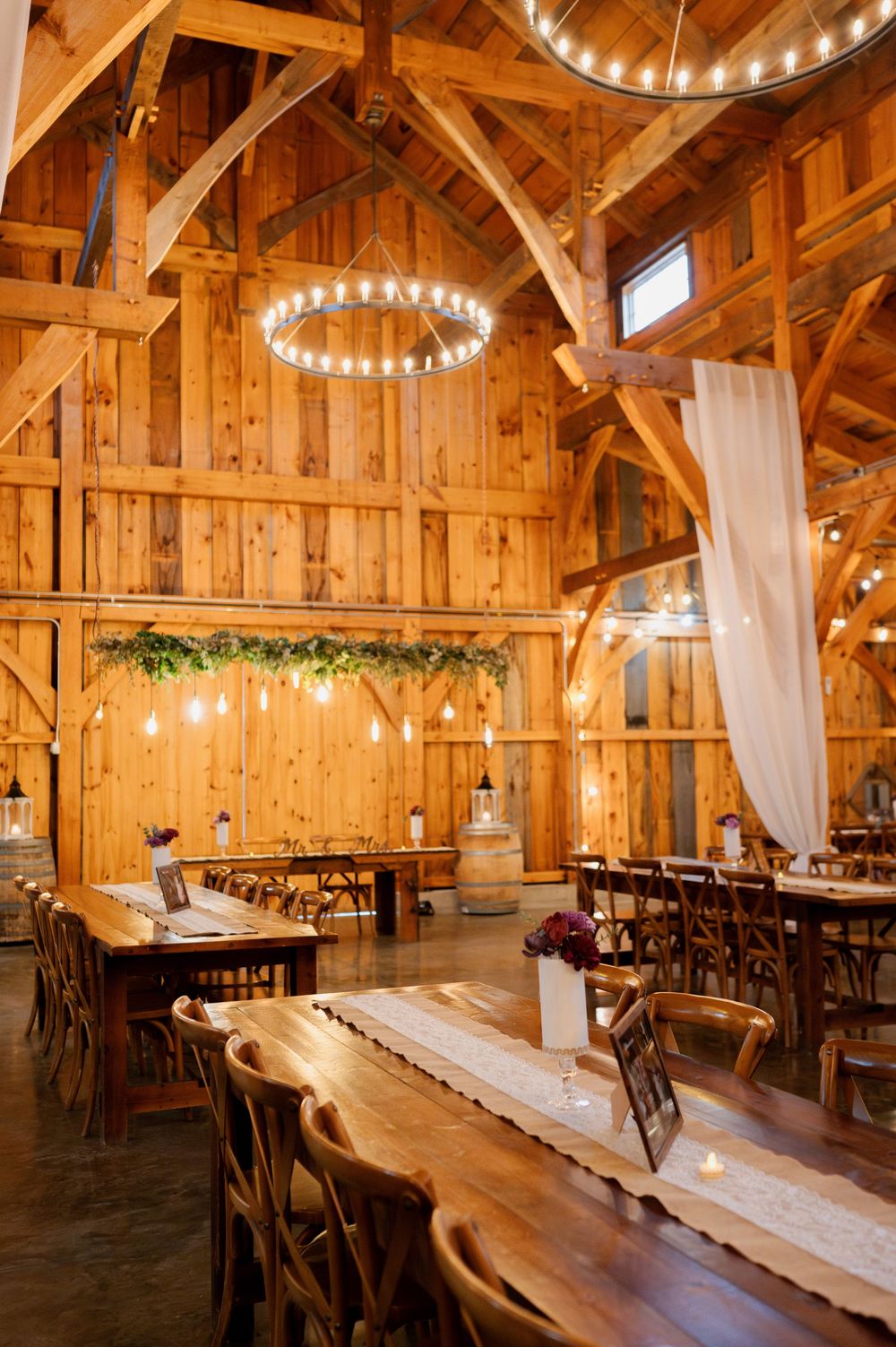 Rustic barn wedding venue with wooden beams, string lights, and elegant white drapes creating a warm, romantic atmosphere.