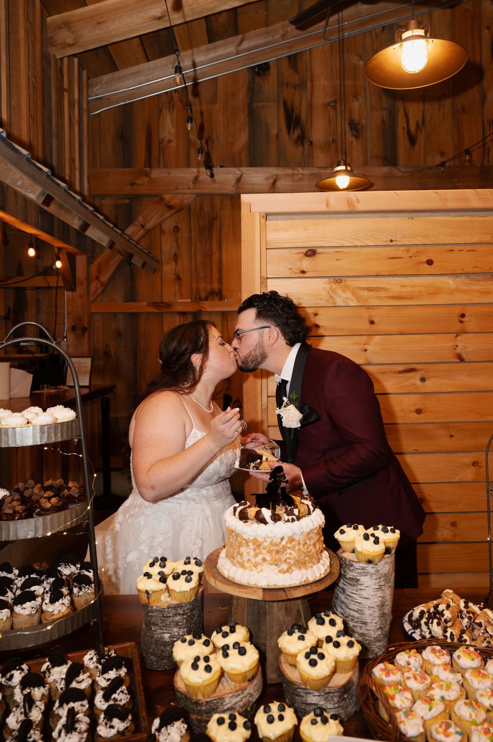 A rustic wooden barn wedding reception features an elegant dessert display with tiers of cupcakes and cake.