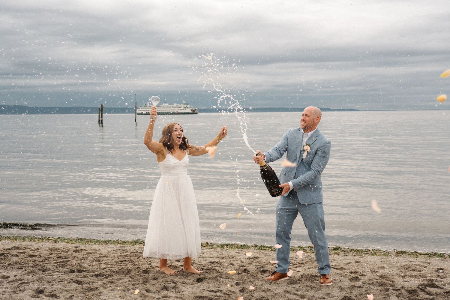 Celebratory moment on a beach with champagne spray against cloudy skies and ocean backdrop.