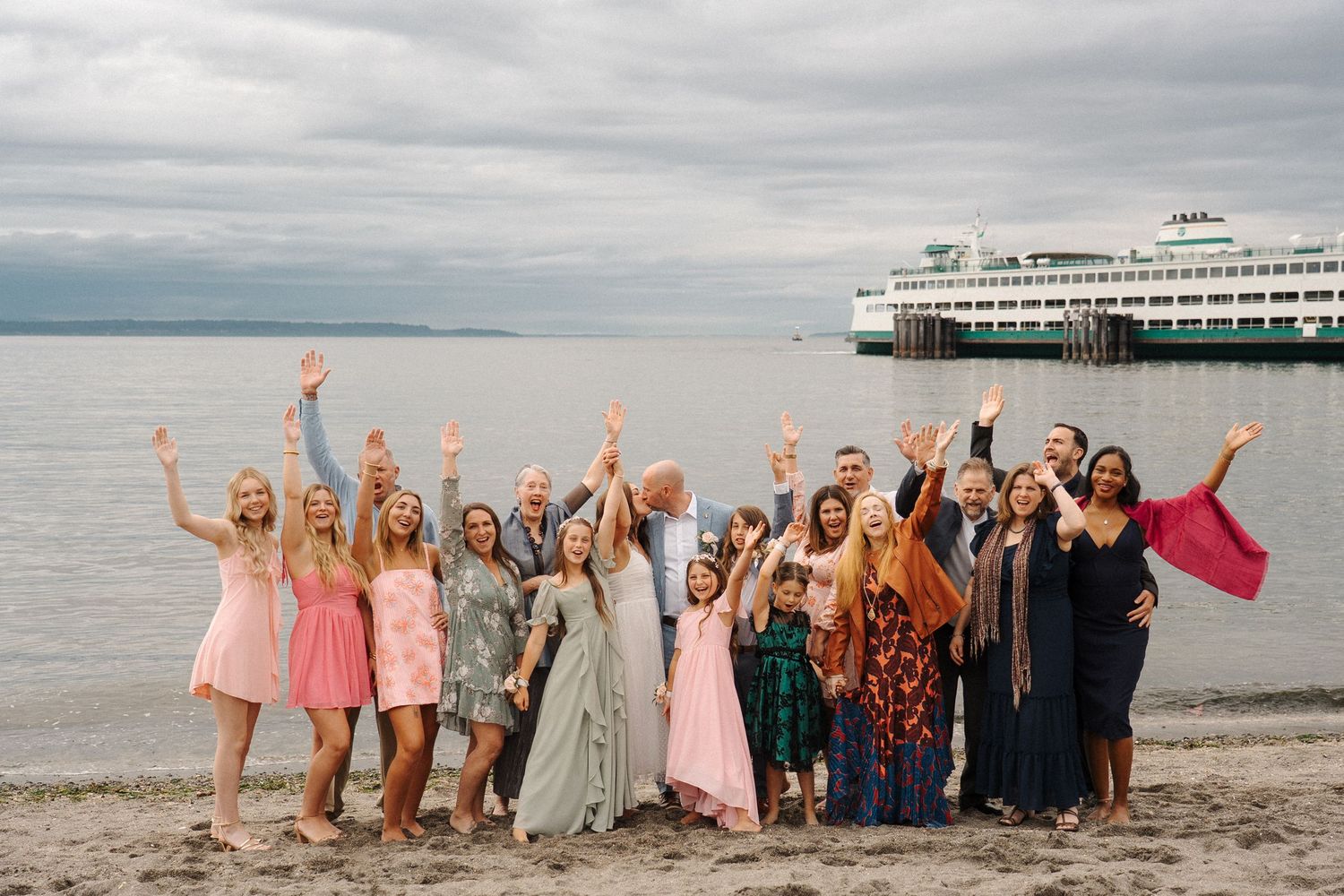 A group of people celebrate joyfully on the beach with raised arms as a large ferry boat passes in the background.