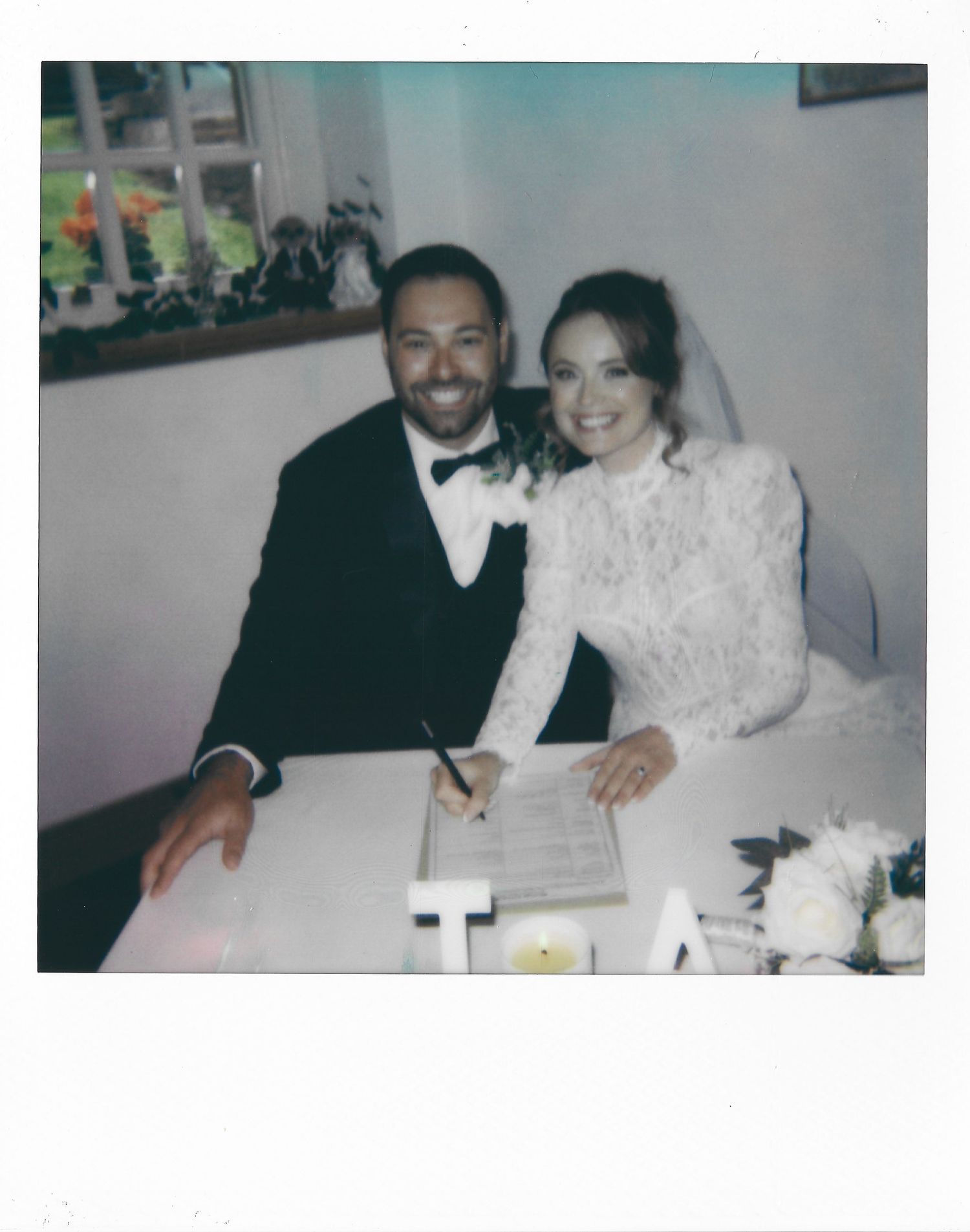 A couple in formal wedding attire signs paperwork at a table with white floral decorations.