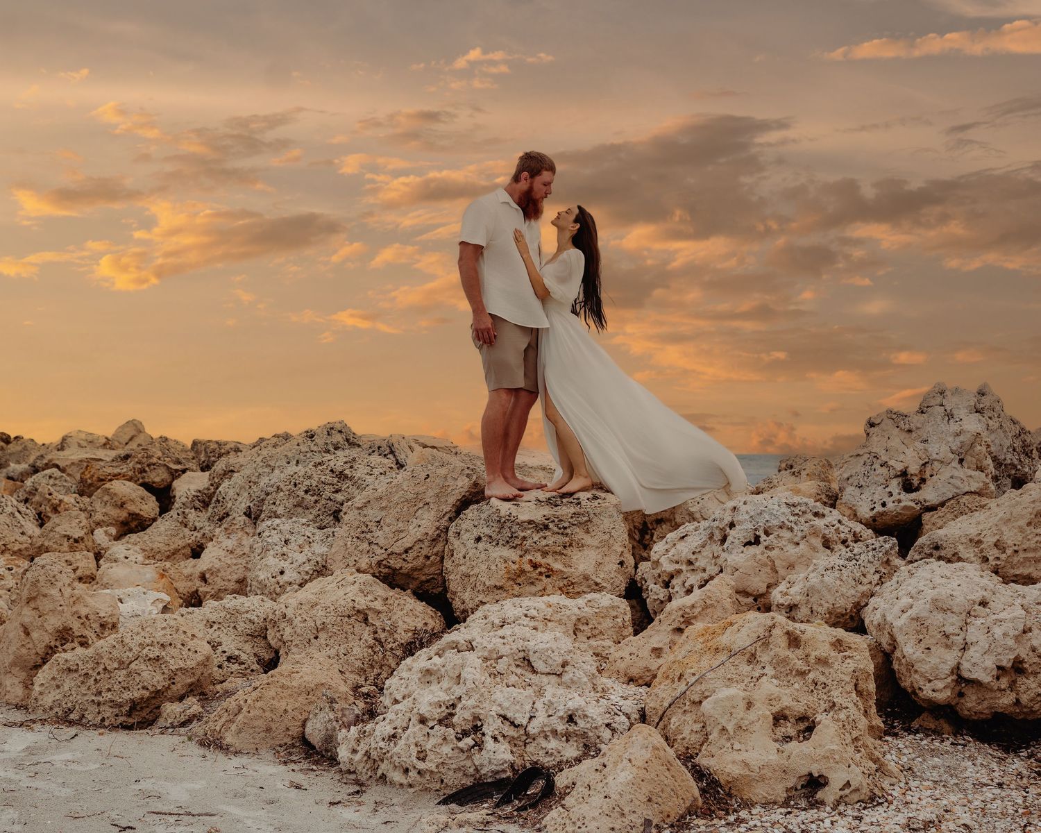 A couple in elegant attire poses on coastal rocks during a dramatic orange and pink sunset.