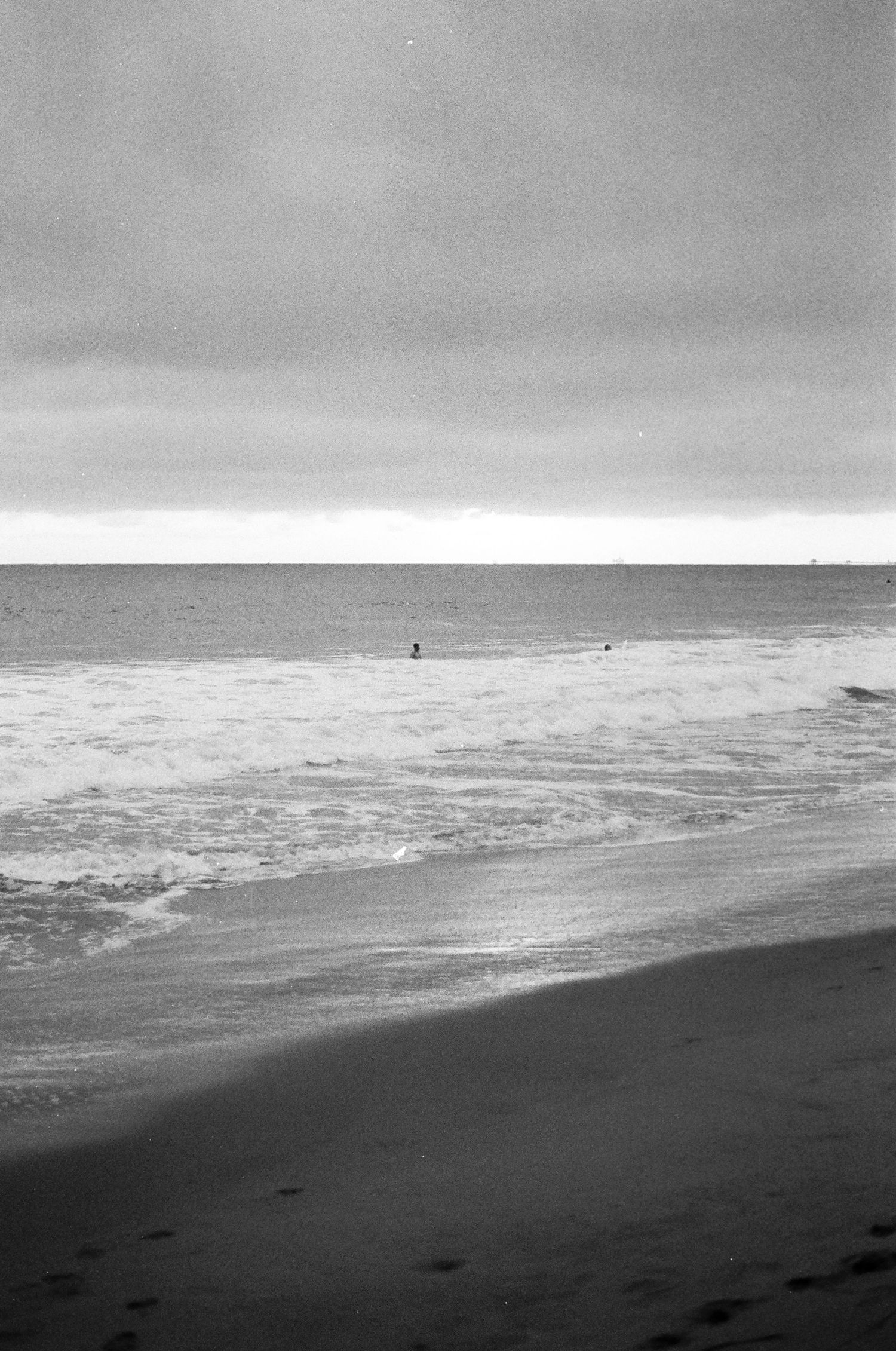 Grayscale seascape panorama shows rolling waves meeting a dark sandy beach under an overcast sky.