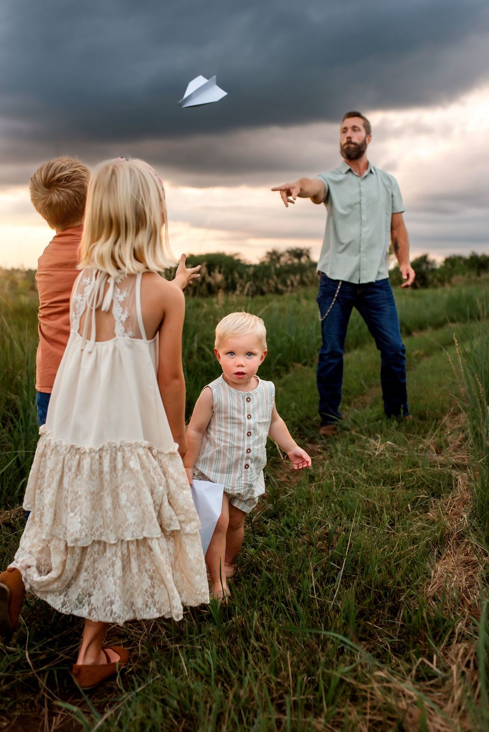 Family playing with paper airplanes in a grassy field under dramatic stormy clouds at sunset.