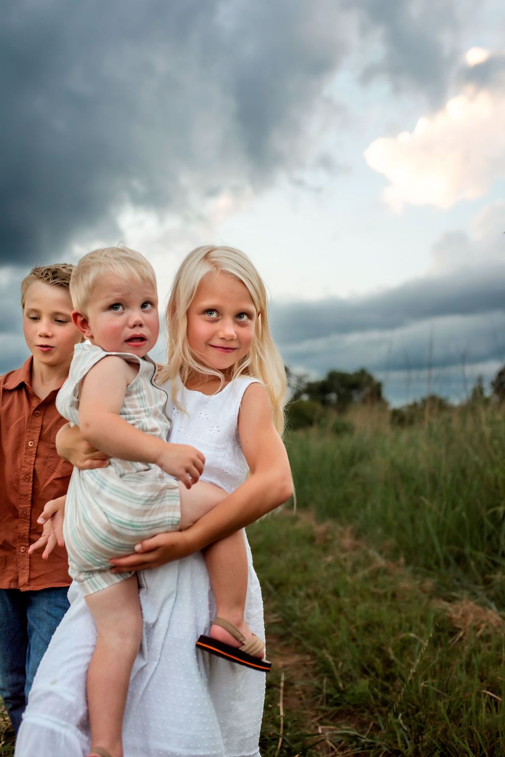 Three children standing together in a grassy field under dramatic stormy clouds at dusk.