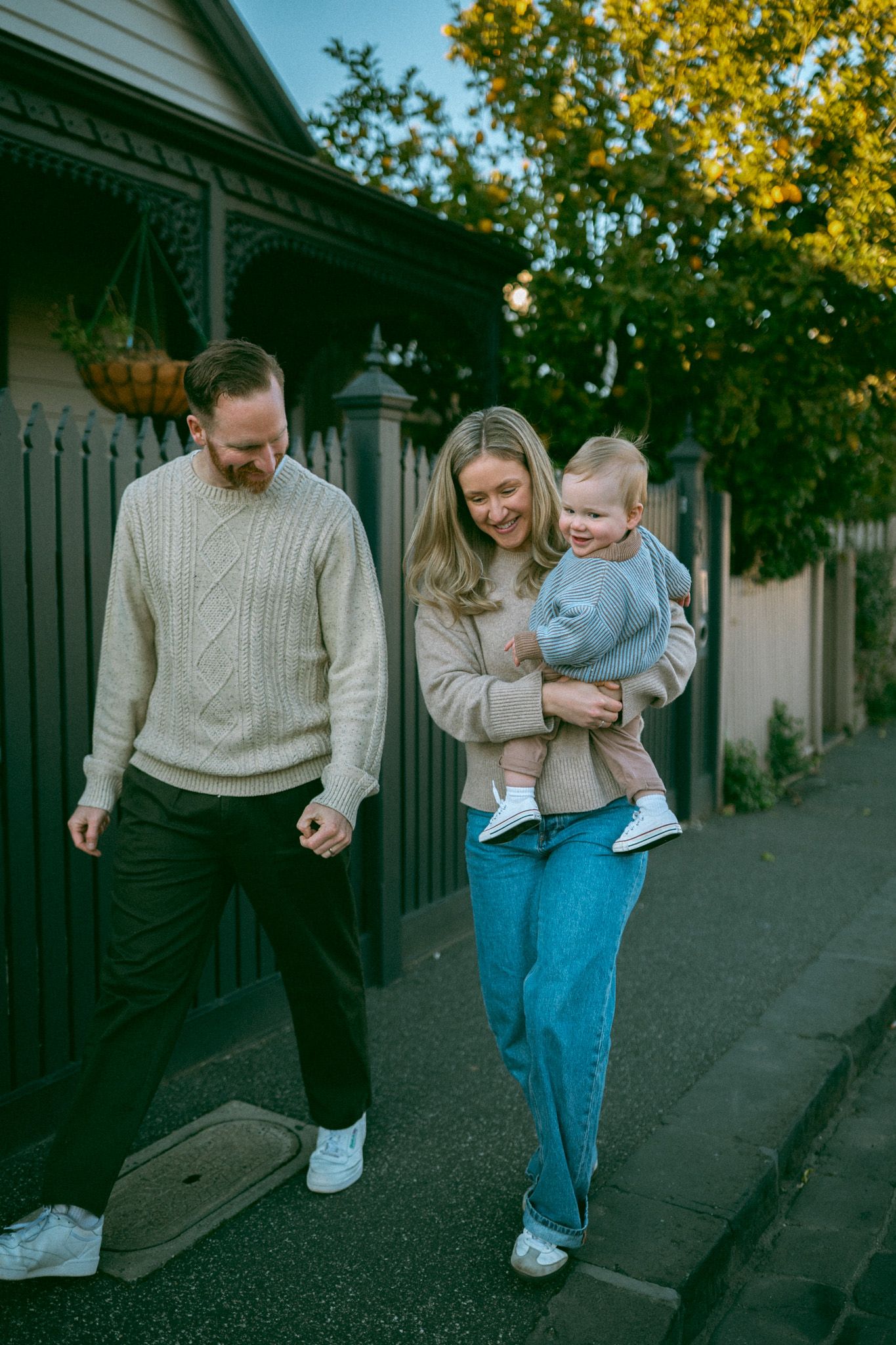 A couple in casual sweaters and jeans walks together on a street with fall trees in the background.