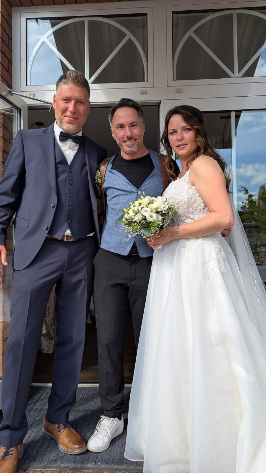 Traditional wedding photo with people posing outdoors in front of ornate doorway with white dress and bouquet.