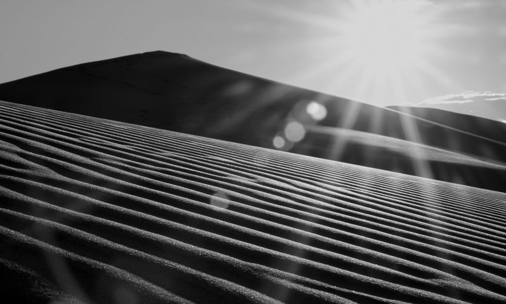 Black and white sand dune with sun rays bursting over the ridge creating lens flare.