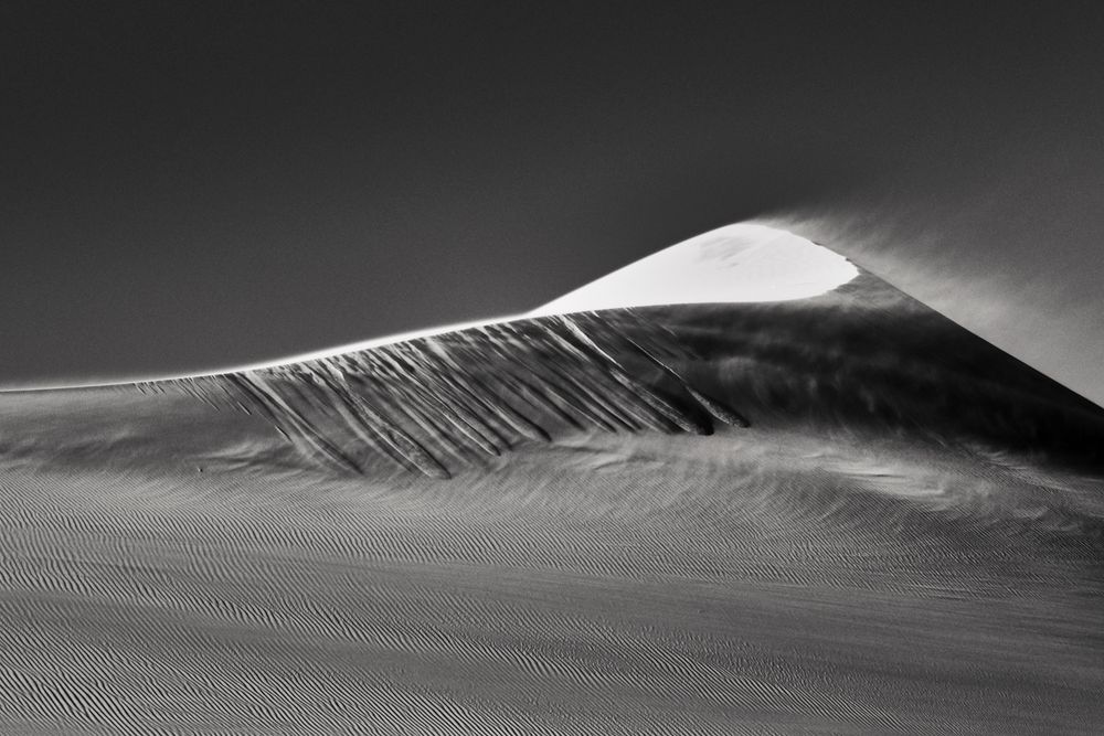 Black and white dramatic sand dune peak with wind-swept ridges against a clear sky.