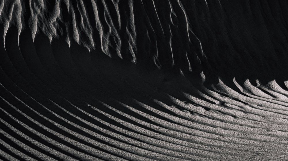 Black and white aerial view of sand dune ridges creating dramatic rippling texture patterns.