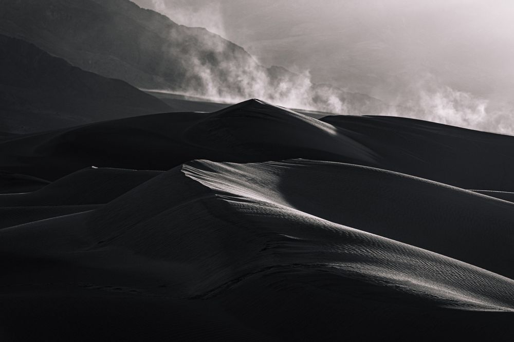 Dark moody black and white sand dunes with dramatic clouds and misty mountains in background.