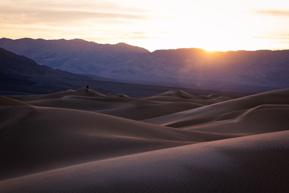 Stunning sunset over rolling sand dunes with silhouetted mountains and lone figure in desert.